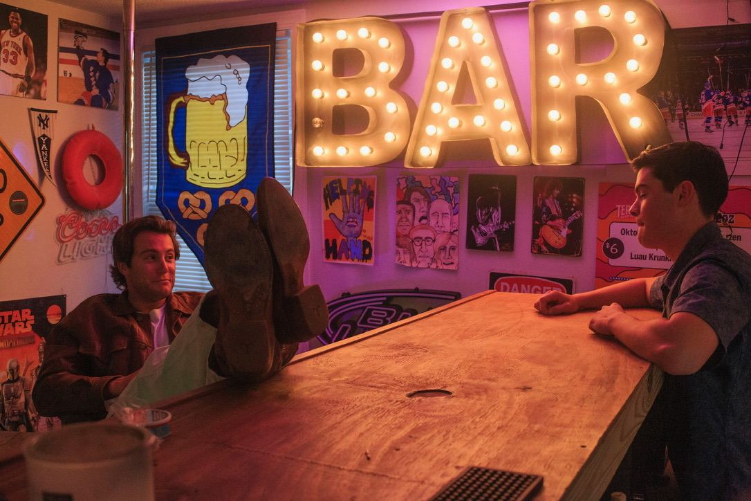 Dante with his boots up on a wooden bar under a lit BAR sign, talking with a friend