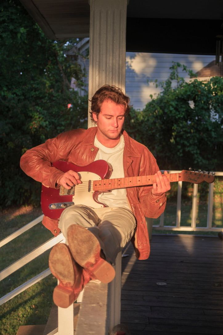 Dante playing a red electric guitar on a porch with his boots up on the railing
