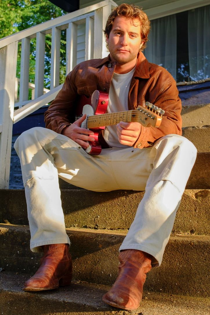 Dante sitting on concrete steps with a red electric guitar, in a leather jacket and boots