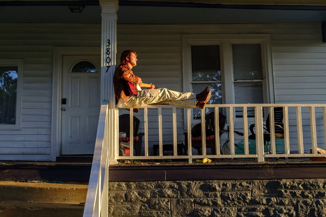 Dante sitting on a porch railing with a guitar, lit by low evening sun