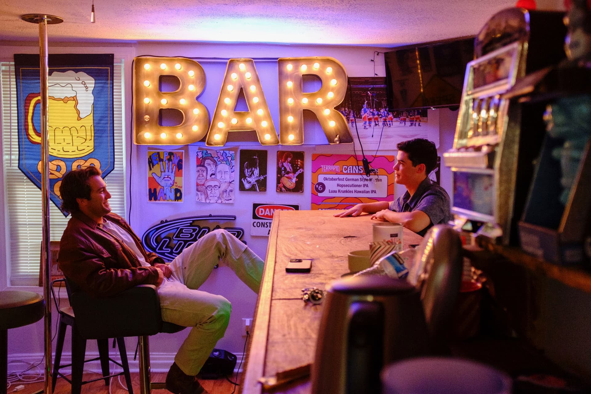 Dante DeRosa laughing at a bar with a friend under a vintage marquee BAR sign