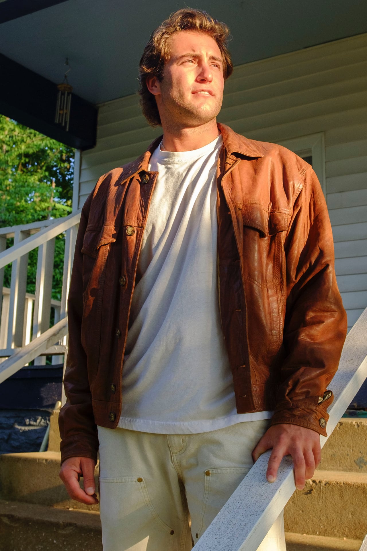 Dante DeRosa standing on the porch steps in a worn leather jacket, lit by warm evening sun