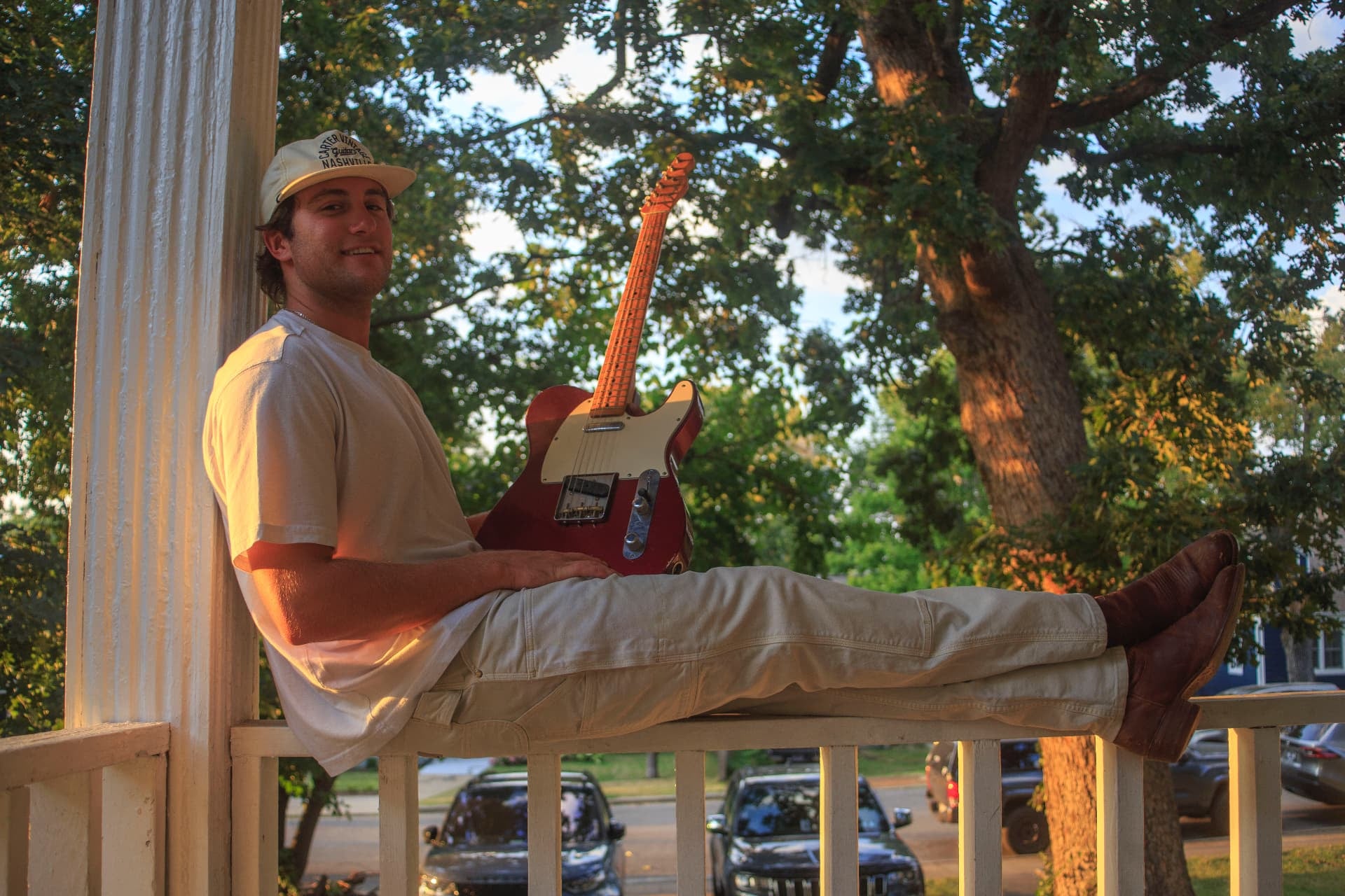 Dante sitting on a porch railing with a guitar, lit by low evening sun